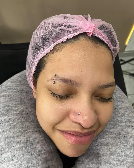 Cliente sorrindo, de touca descartável, com piercings recém-colocados na sobrancelha e no nariz